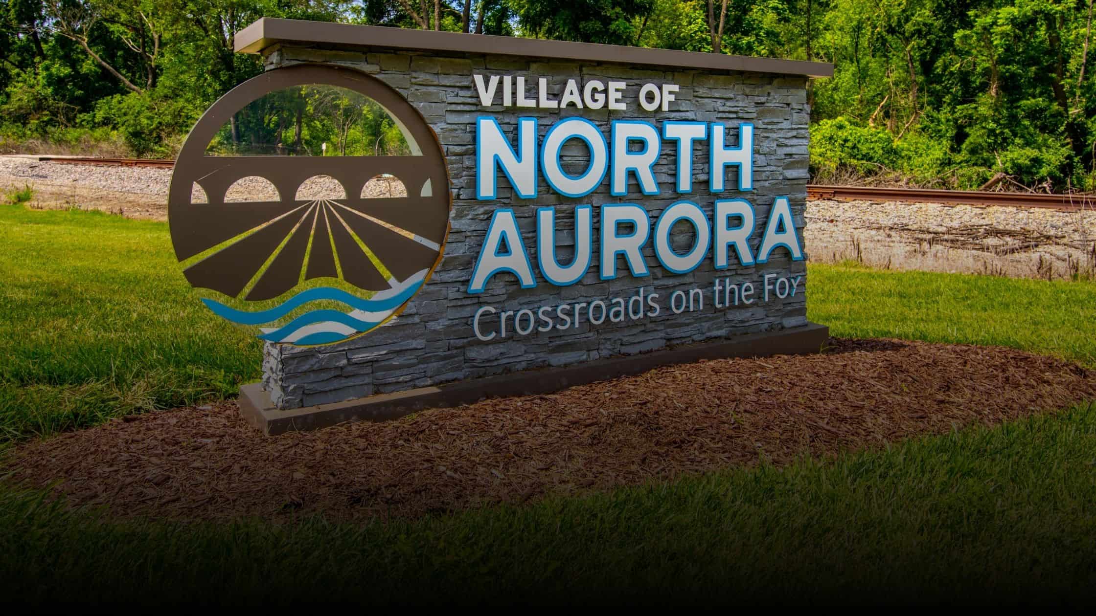 Village of North Aurora welcome sign, reading 'Crossroads on the Fox,' with a scenic bridge and river logo